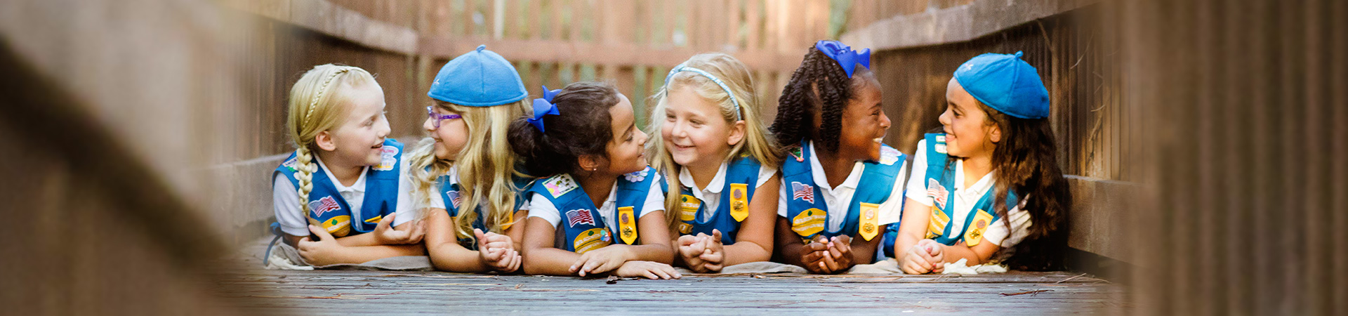  group of daisy girl scouts laying on boardwalk smiling at each other wearing daisy uniform vests, aprons, and hats. 